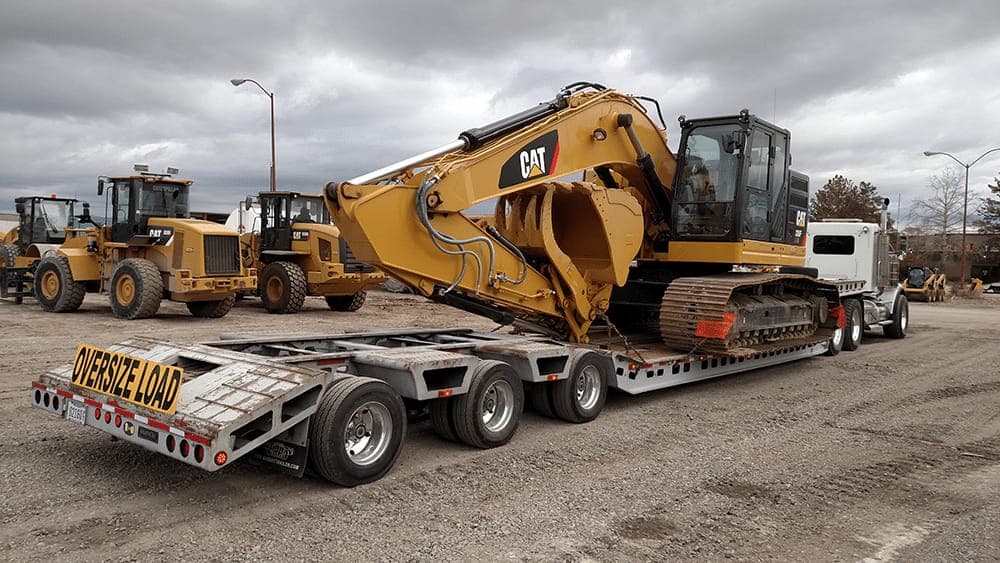 CAT excavator loaded on a lowbed trailer marked "Oversize Load", with other heavy equipment in the yard
